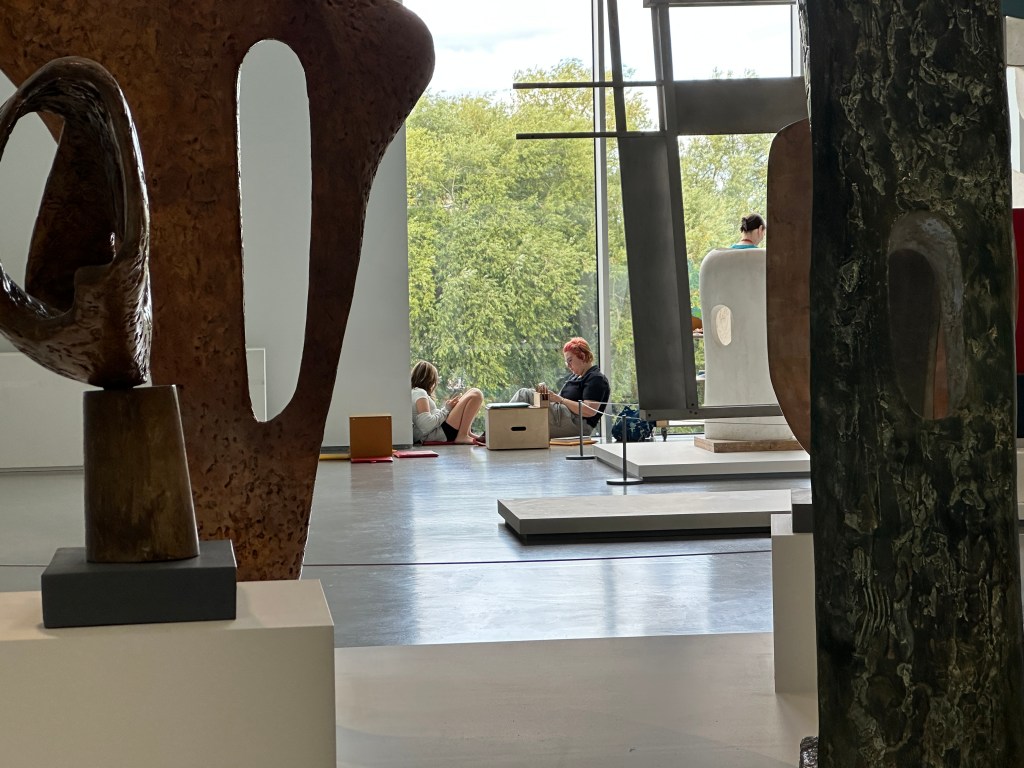 Interior view of an art gallery featuring sculptures, with visitors seated and engaging with the artworks in the background and natural light coming in through large windows.